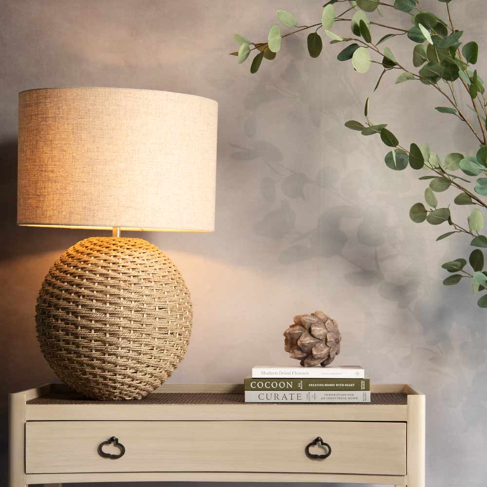 A woven rattan table lamp with a cylindrical beige fabric shade sits on a light wood side table, casting a warm glow. The table has black metal drawer pulls and holds books and a decorative object, set against a textured gray wall with eucalyptus branches hanging from the top right.