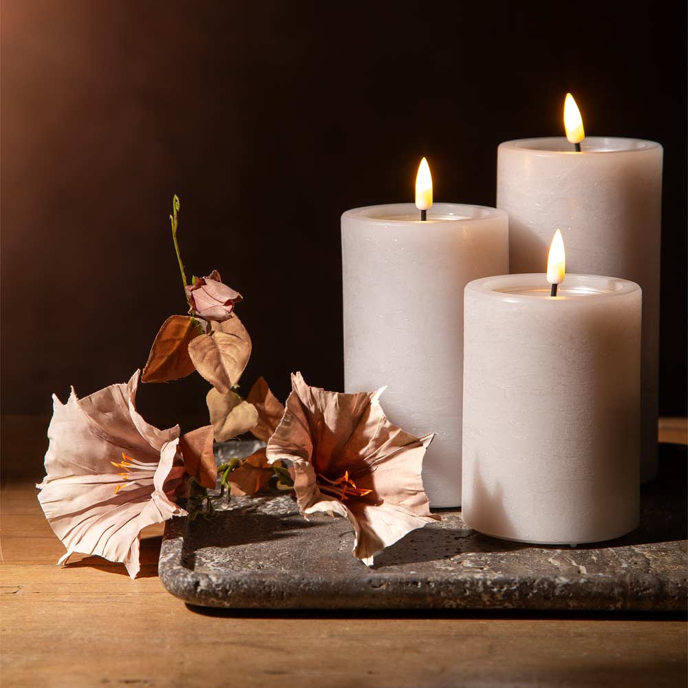 Three lit matte pillar candles in a soft blush pink colour, arranged on a brown marble tray on a rustic wooden tabletop with  a faux flower stem alongside.