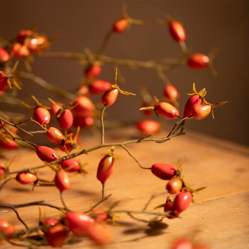 Artificial rosehip branch garland with small red-orange berries on thin brown stems, arranged on a wooden surface against a warm brown background.