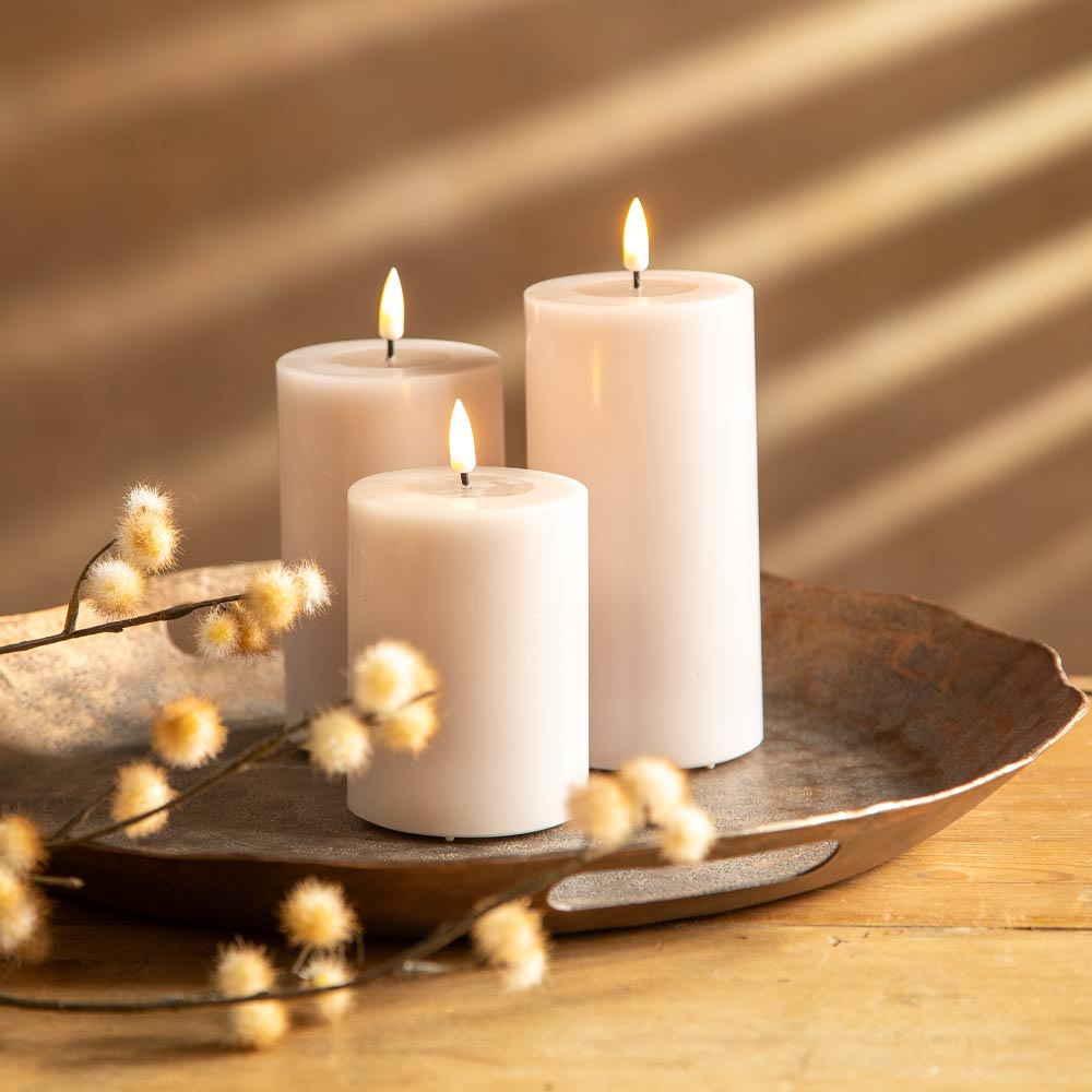 Three lit rose pillar candles arranged on a rustic metal tray with faux flowers, set on a wooden surface in warm sunlight with diagonal shadows.