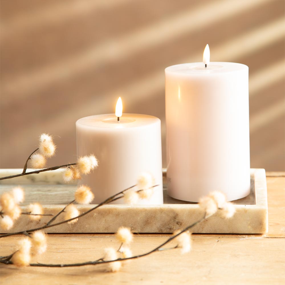 Two lit rose pillar candles arranged on a marbel tray with faux pom pom styled flowers, set on a wooden surface in warm sunlight with diagonal shadows from a window blind.