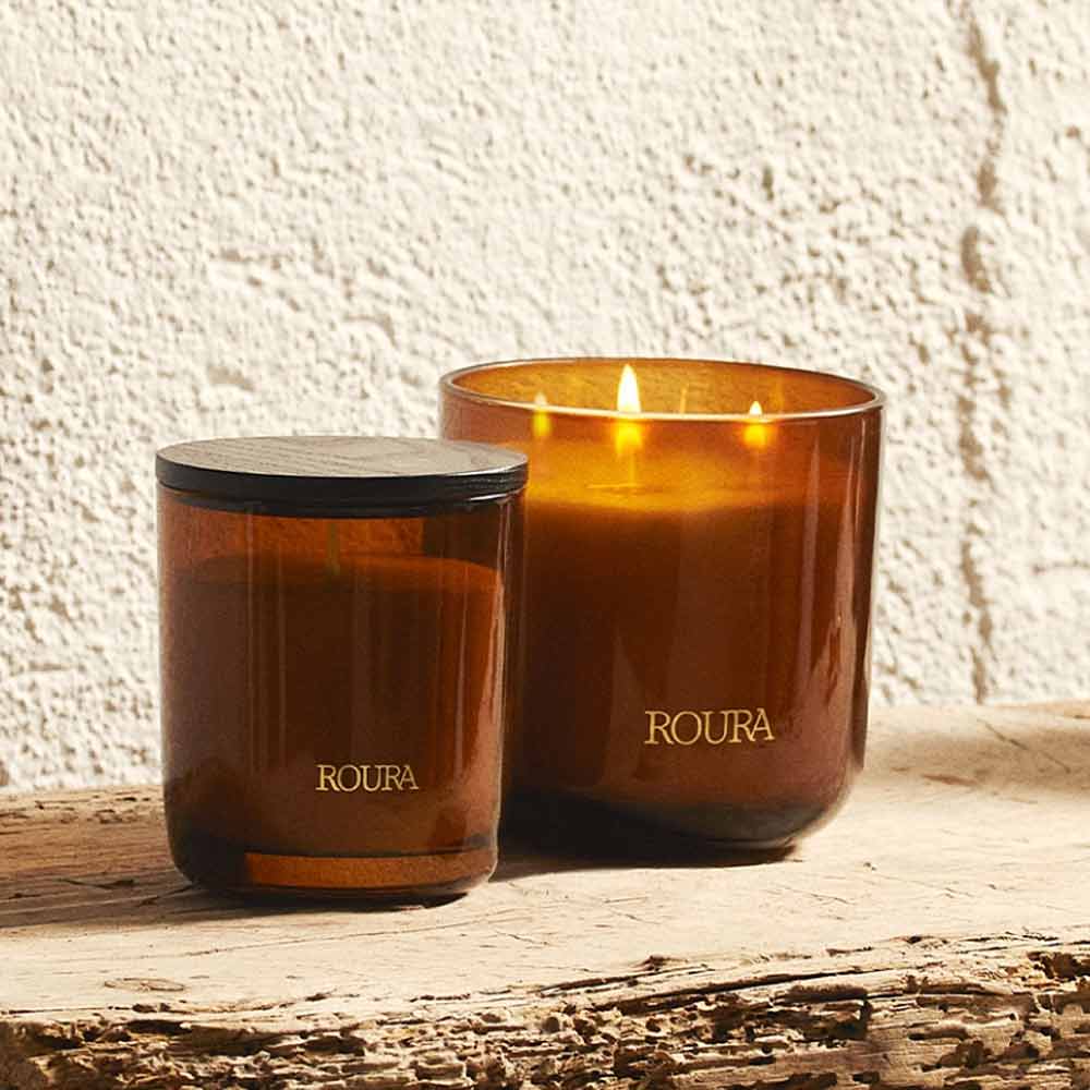 Two ROURA candles on a wooden surface with a textured wall background includes Royal Amber 3 Wick Candle by Roura