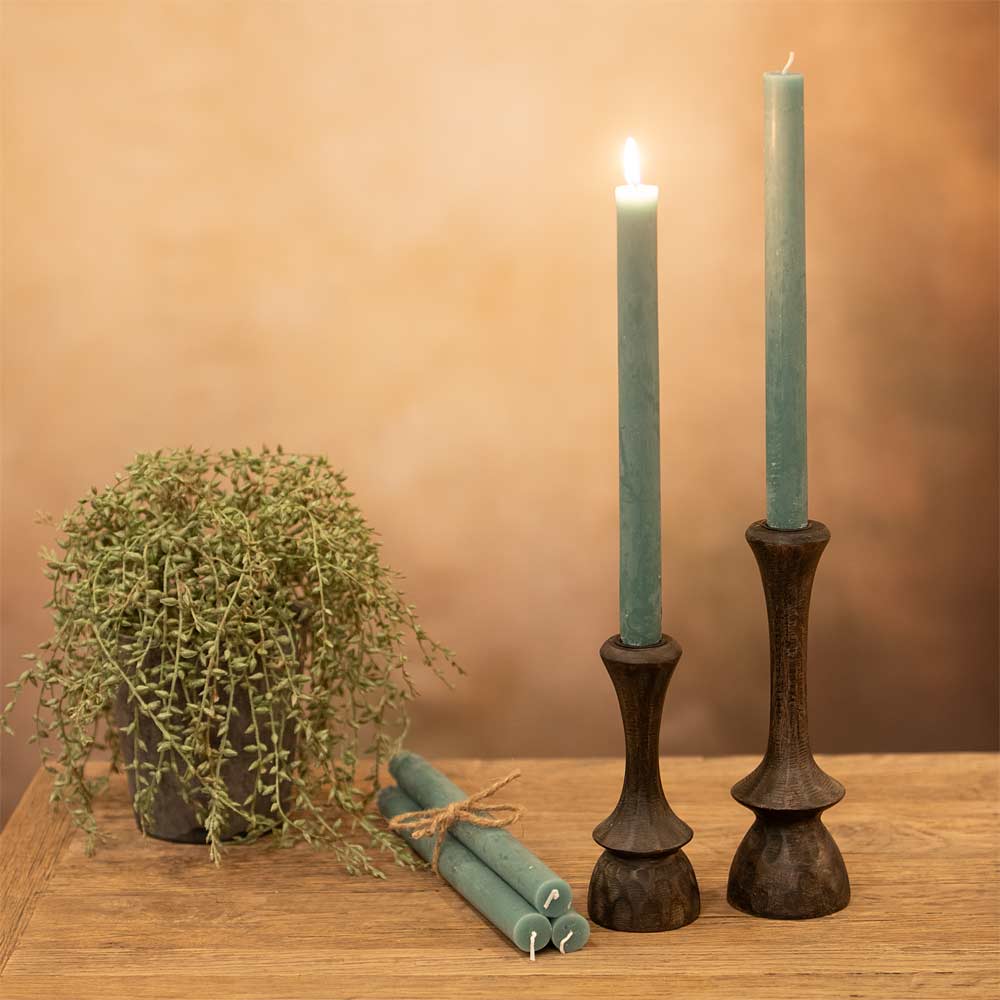 Two Sea Green candles in rustic candlesticks on a wooden surface with a plant in the background.