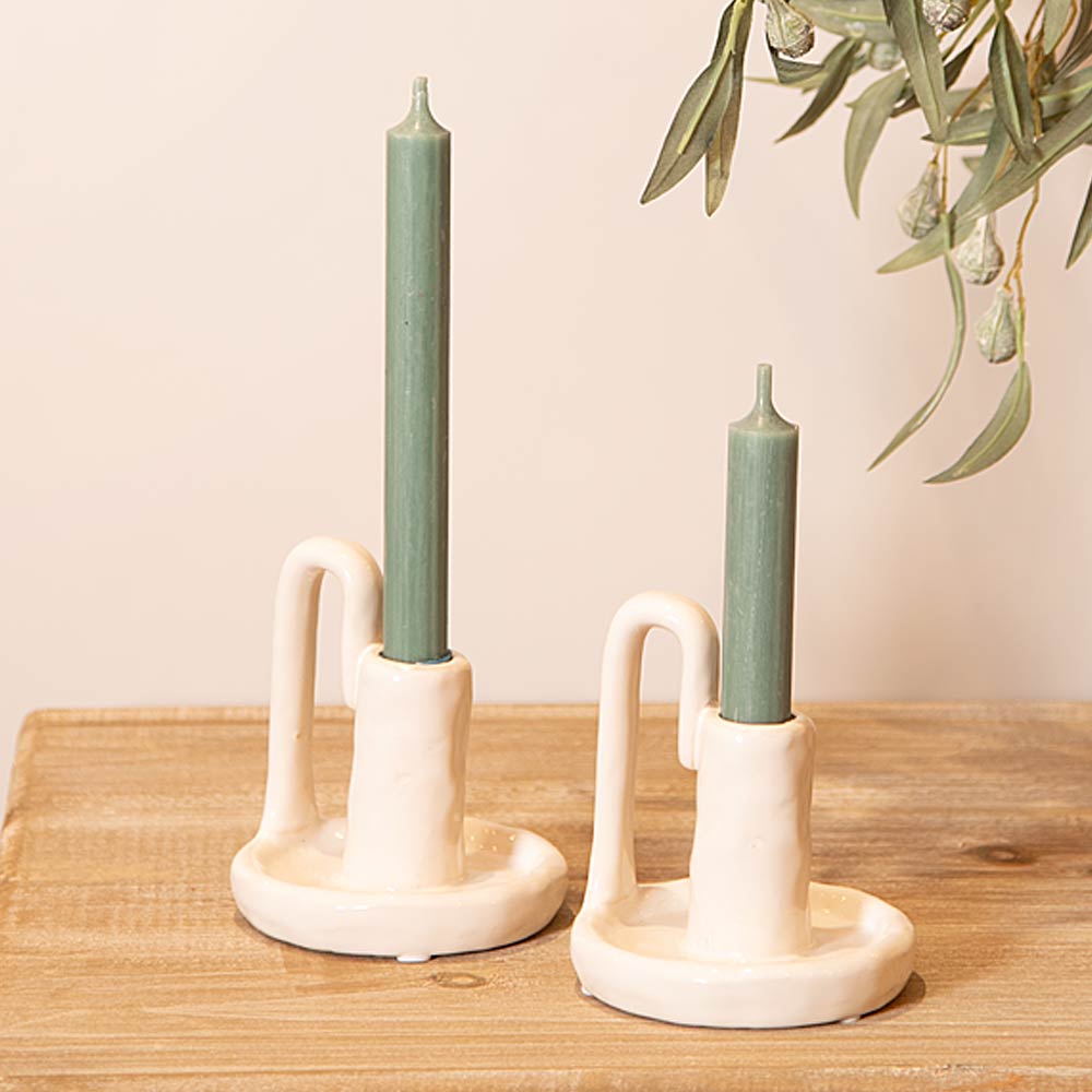Two White Rustic Ceramic Candlesticks with green candles on a wooden surface, with a plant in the background.