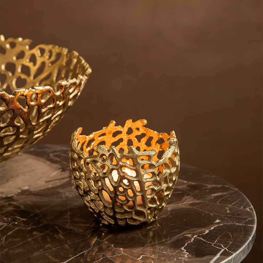 A small gold-toned decorative coral effect candle holder or bowl with a warm glowing candle inside, placed on a dark marble surface against a brown background. A larger matching Bowl shaped piece is partially visible on the rear left.