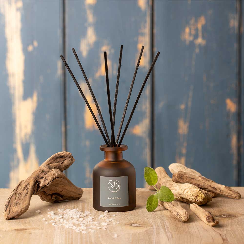 Sea Salt and Sage Reed diffuser with sticks in a brown bottle on a wooden surface with driftwood and sage leaves.