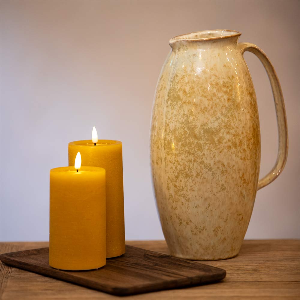 Ceramic pitcher on a wooden surface with two ochre pillar candles alongside in front of a neutral background