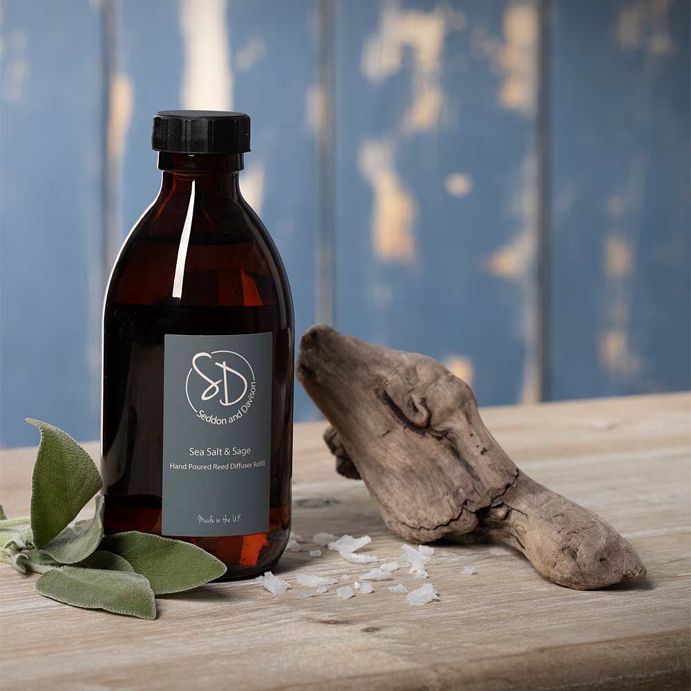 A dark amber glass bottle of Sea Salt & Sage diffuser refill with a black cap and gray label, styled on a wooden surface with sea salt crystals, green sage leaves, and driftwood against a blue wooden background.