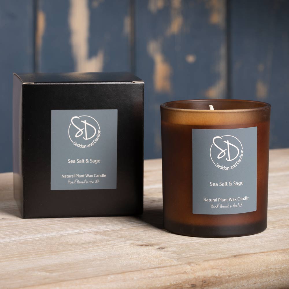 An amber glass votive candle next to its black box packaging, both featuring a grey label for 'Sea Salt & Sage' scent, by Seddon and Davison both items are set on a wooden surface against a rustic blue wooden background.