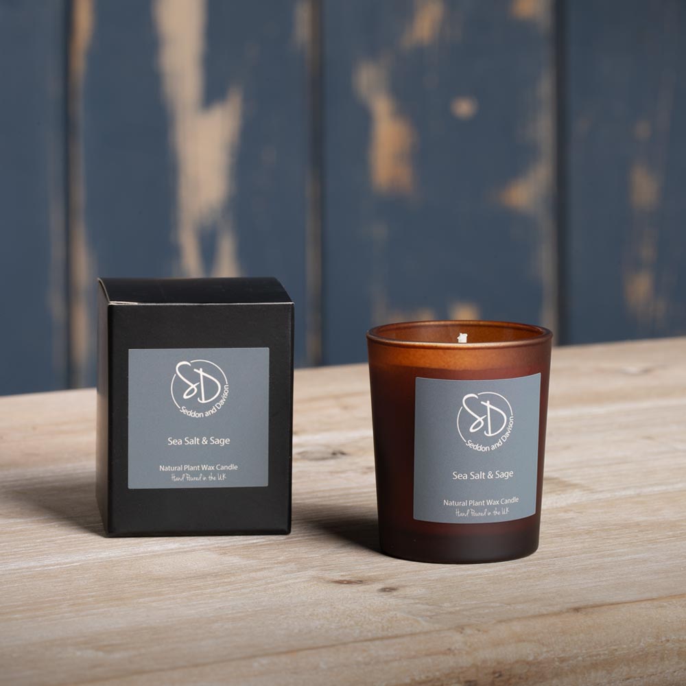 An amber glass Votive candle and its black packaging box, both labeled 'Sea Salt & Sage' by the brand 'Seddon and Davison, set on a light wooden table against a rustic blue wooden wall background.