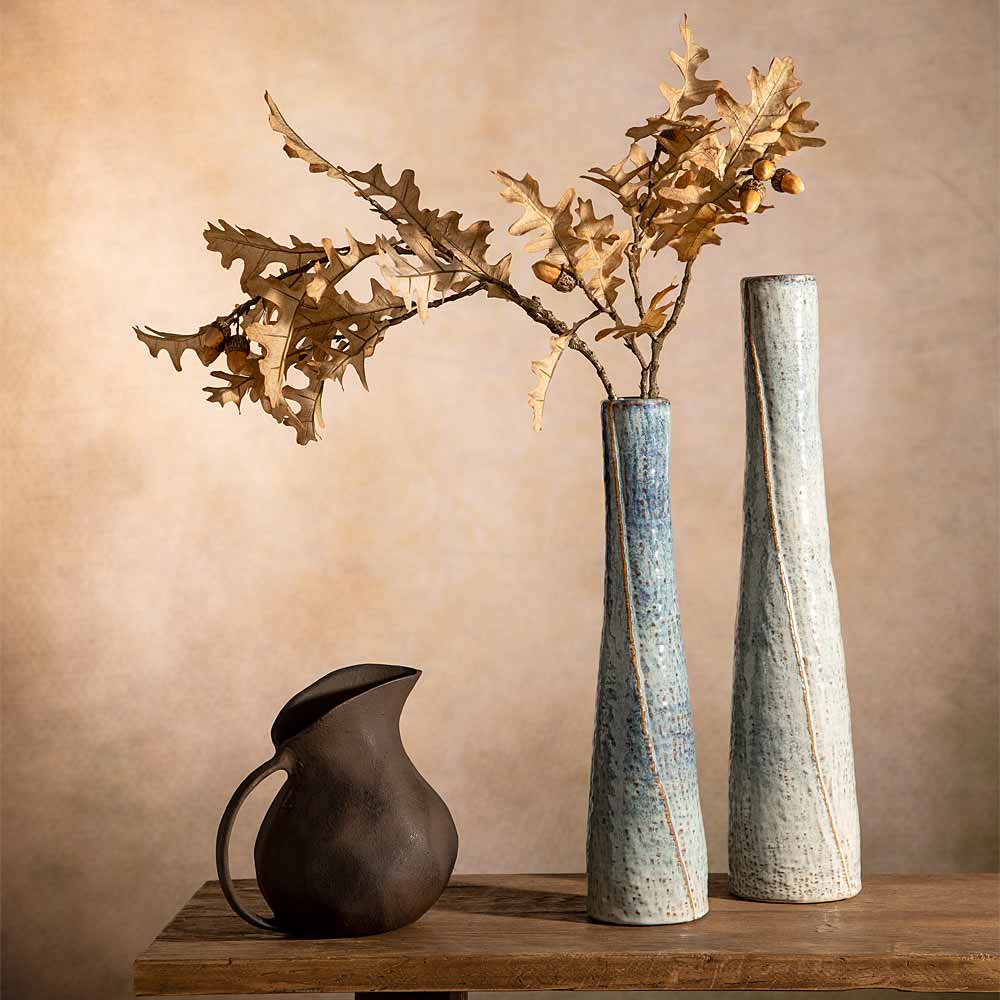 Two tall textured ceramic vases in blue and brown tones with a rustic dark brown pitcher, styled with faux  oak branches, leaves and acorns on a wooden table against a warm beige background.