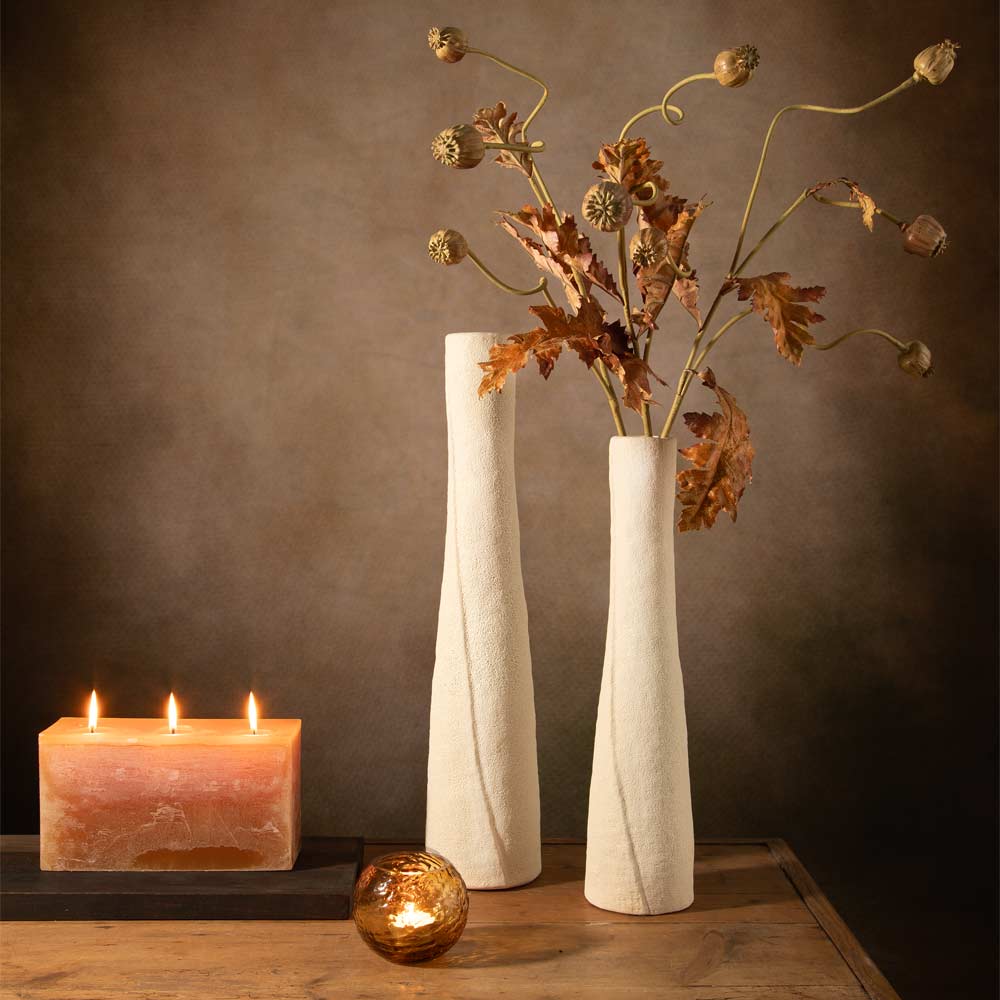 Two tall textured linen white ceramic vases filled with faux autumn leaves and seed pods, displayed on a wooden table next to a lit caramel coloured oblong shaped candle and a small amber glass candle holder against a dark brown background.