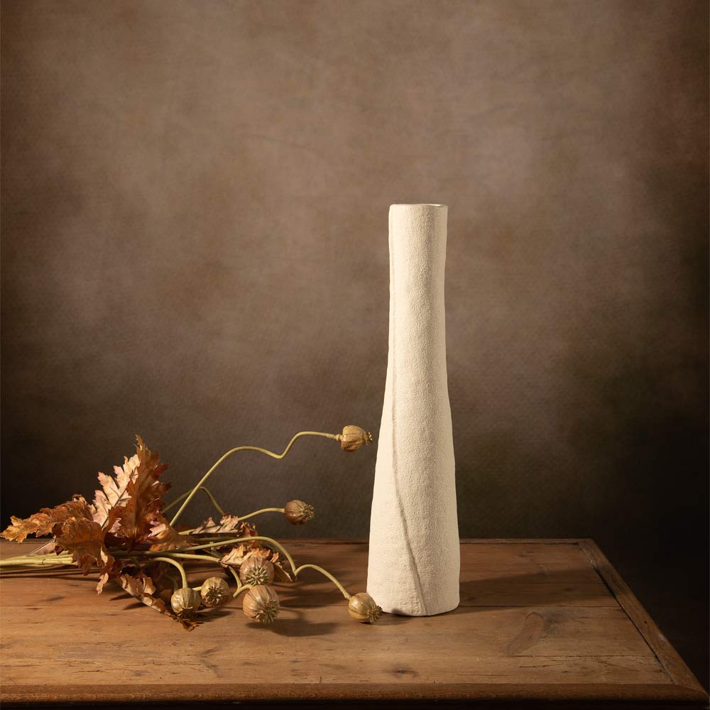 A tall, flared ceramic vase in a textured off-white finish stands on a rustic wooden table against a dark brown  wall. Dried autumn leaves and seed pods are arranged to the left as styling props.