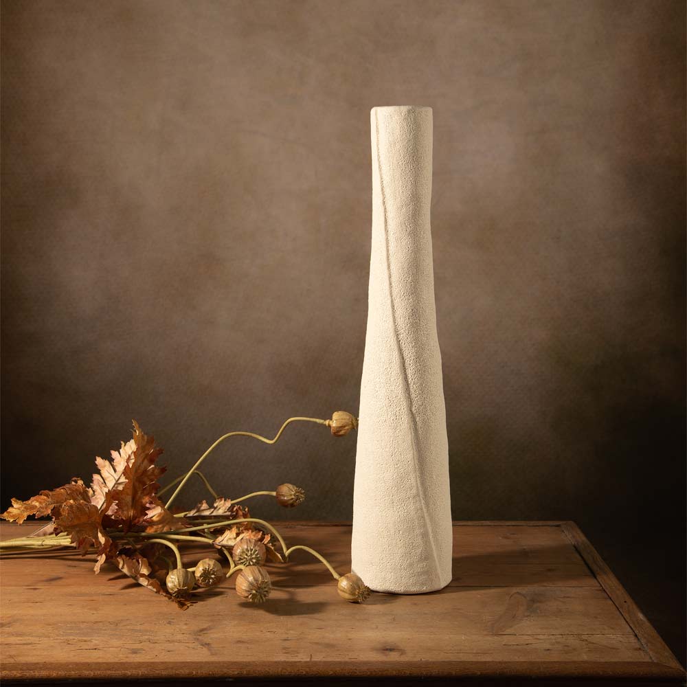 A tall, slender off-white linen coloured ceramic vase with a textured surface and a twisted spiral design, displayed on a rustic wooden table next to faux  botanical stems against a dark brown textured background.
