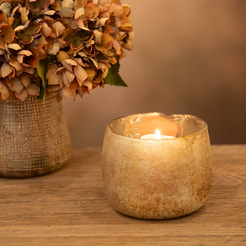 Candle in a textured holder next to a vase of dried flowers on a wooden surface.