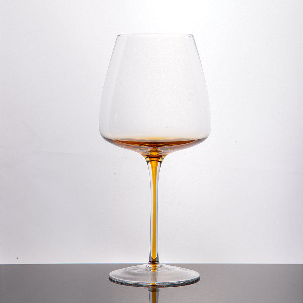 A clear glass wine glass with a wide bowl that grades to and amber base leading down the stem amber slender stem, shown on a dark surface against a white background.