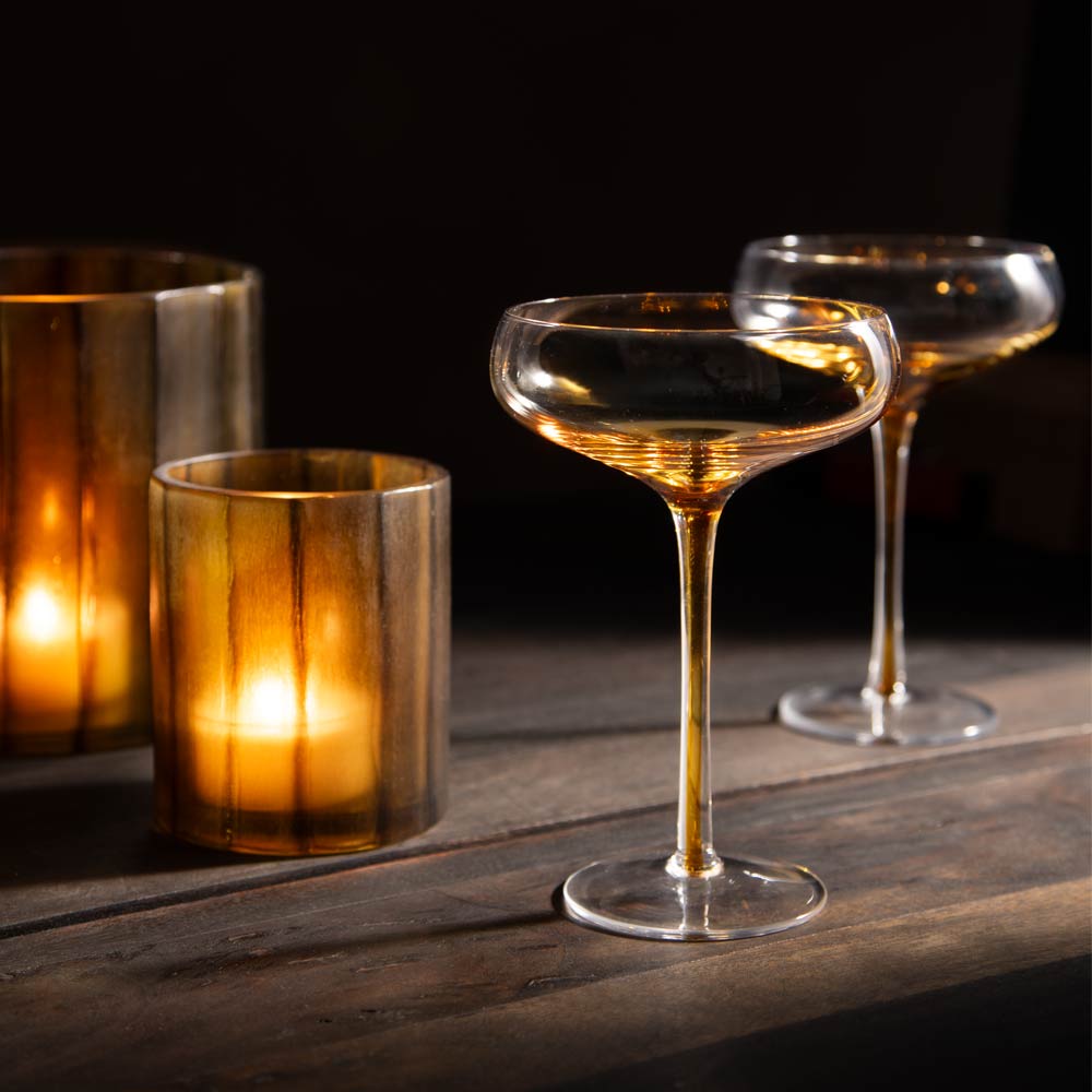 A clear stemmed coupe-style glass with an amber gradient towards the base of bowl into the stem, shown on a dark wooden surface beside lit candle holders. 