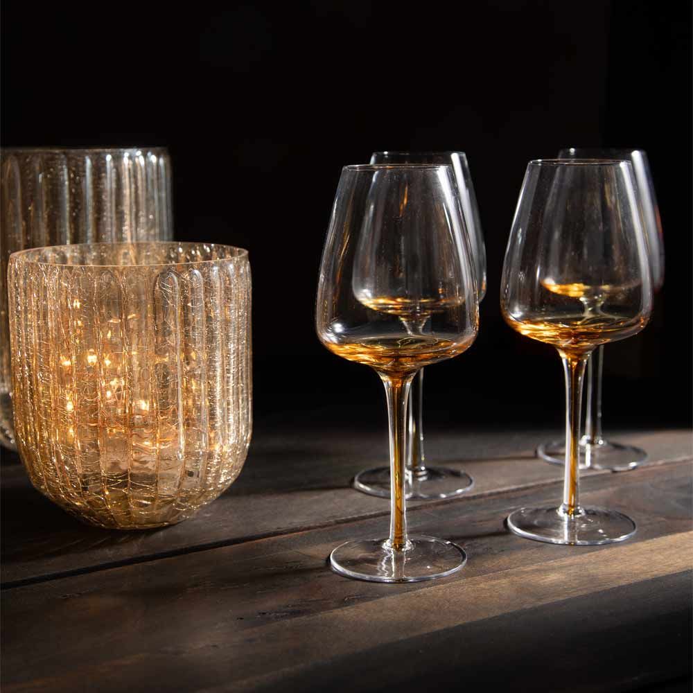 Four clear stemmed wine glasses with a wide bowl that is clear and has a gradient of amber towards the base and  into the stem , arranged on a dark wooden surface against a black background, with a lit ribbed glass candle holder beside them.