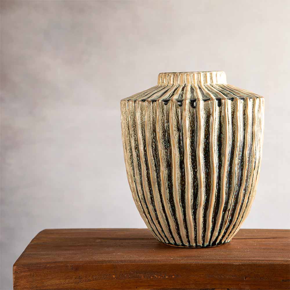 A ribbed ceramic vase with a textured olive and beige finish, displayed on a rustic wooden surface against a neutral gray background.