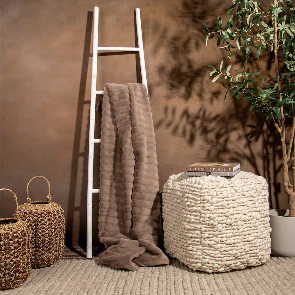 A styled interior scene with a taupe textured throw blanket draped over a white ladder, a chunky cream pouf with books on top, woven baskets, and a potted plant against a warm brown wall.