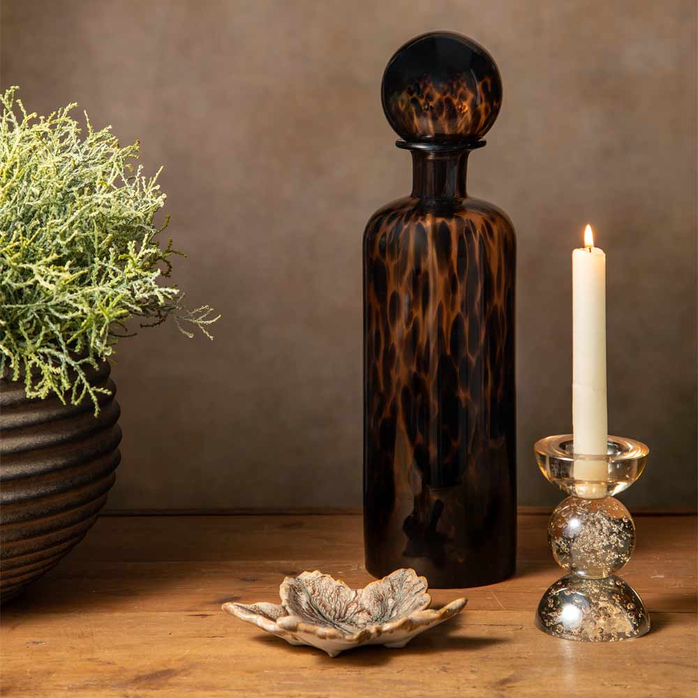 A dark amber-brown glass bottle with a rounded stopper, styled on a wooden surface next to a lit white candle in a Glass holder and a small leaf-shaped dish, with a faux potted plant in the background against a warm brown wall.