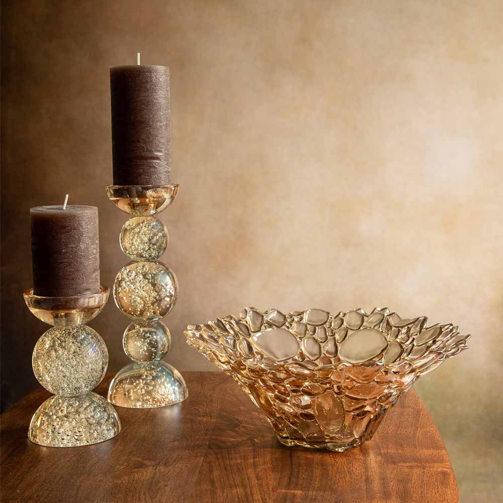 Decorative glass candlestick holders with two dark brown pillar candles and a  textured glass bowl, arranged on a wooden surface against a warm beige background.