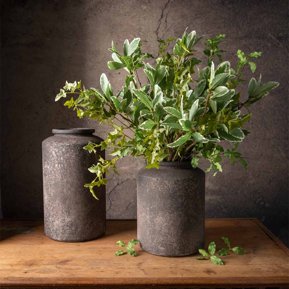 Two rustic and speckled finished black and brown metal cylinder shaped vases, placed on a wooden table, with one vase holding a range of leafy green faux plant stems and the other left empty, set against a textured dark grey wall.