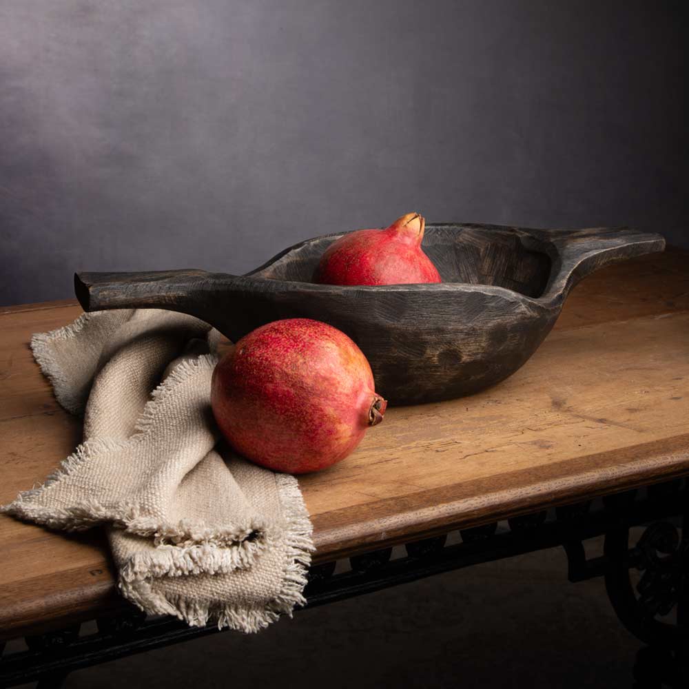 A rustic dark wood serving bowl with two red pomegranates, styled on a wooden table with a beige linen cloth against a dark gray background.