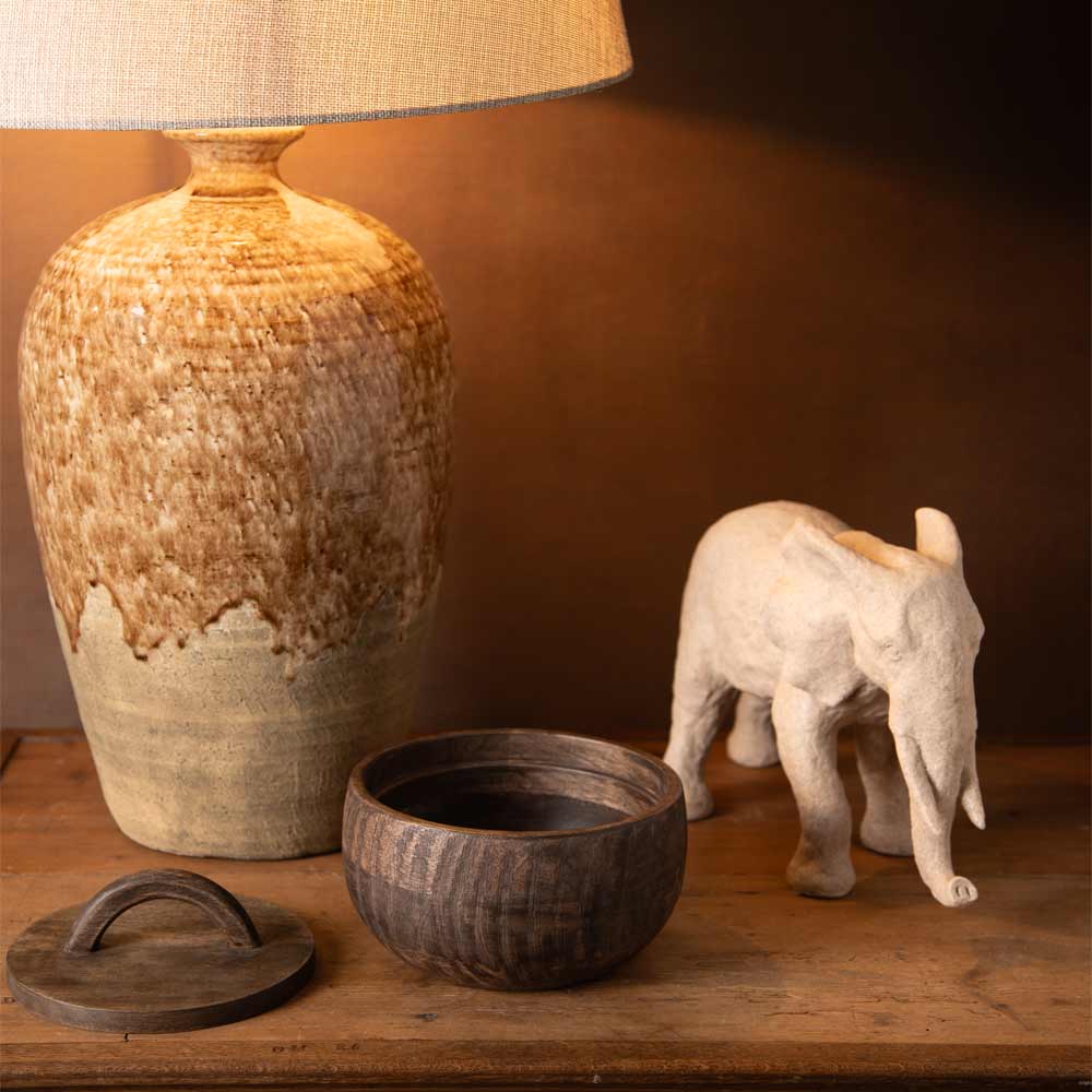 A rustic ceramic table lamp with a textured beige and brown glaze sits on a wooden surface next to a small beige elephant figurine and a dark wooden bowl with its lid.