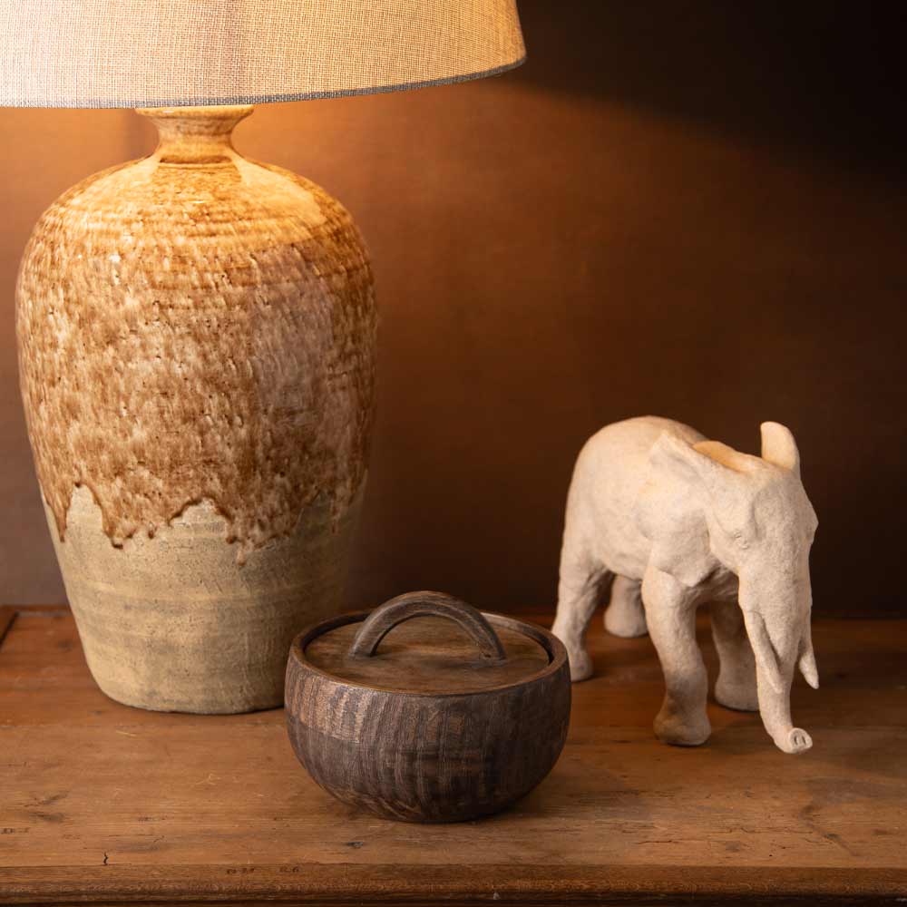 A rustic ceramic table lamp with a textured beige and brown glaze, styled on a wooden surface next to a small elephant figurine and a dark wooden lidded bowl.