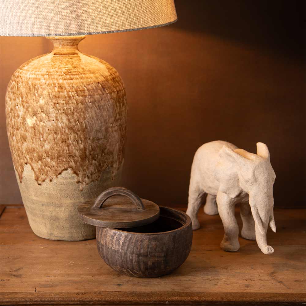 A rustic ceramic table lamp with a textured beige and brown glaze sits on a wooden surface next to a small beige elephant figurine and a dark wooden bowl with its lid on.