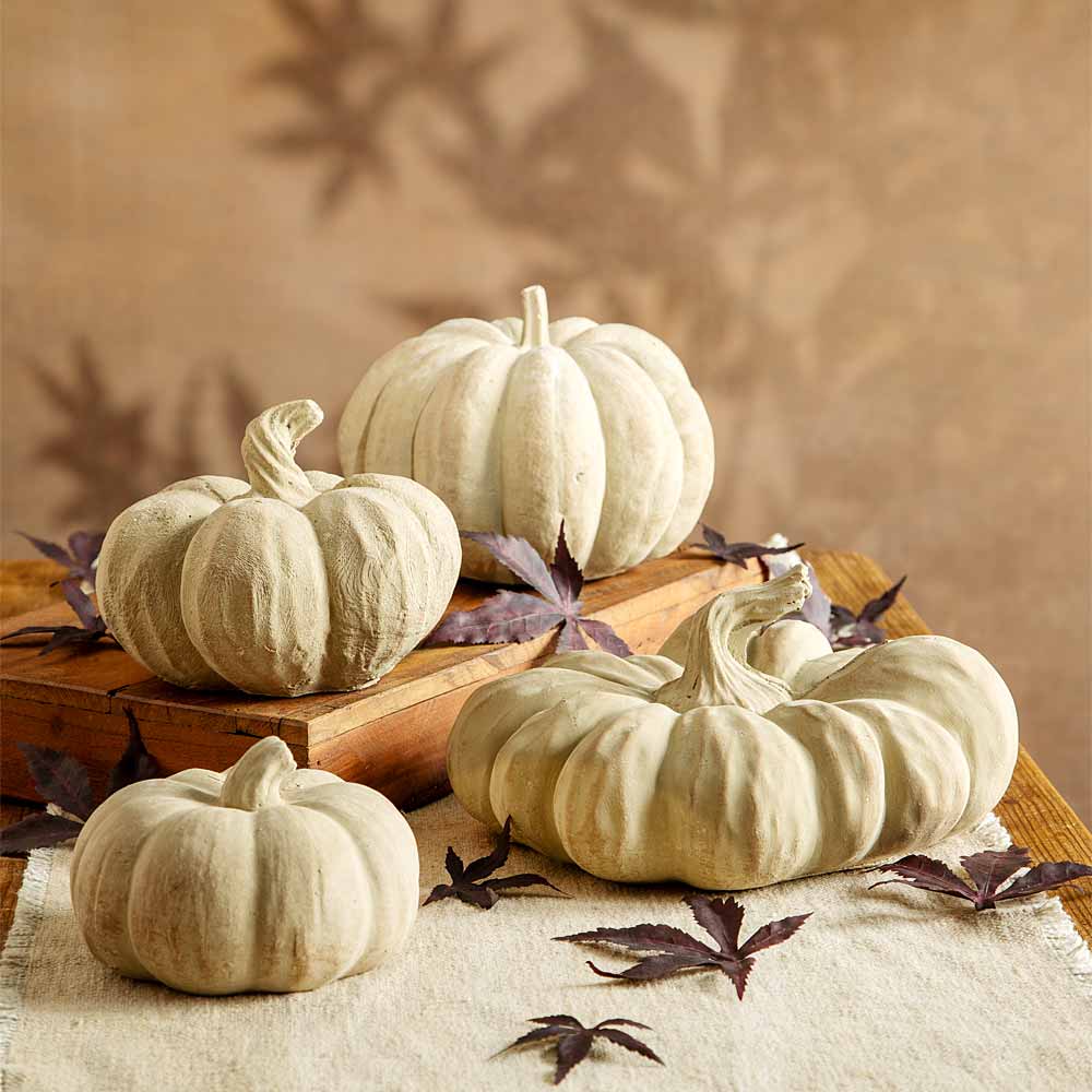 Four decorative faux beige  pumpkins arranged on a rustic wooden surface with scattered dark maple leaves against a warm beige background.