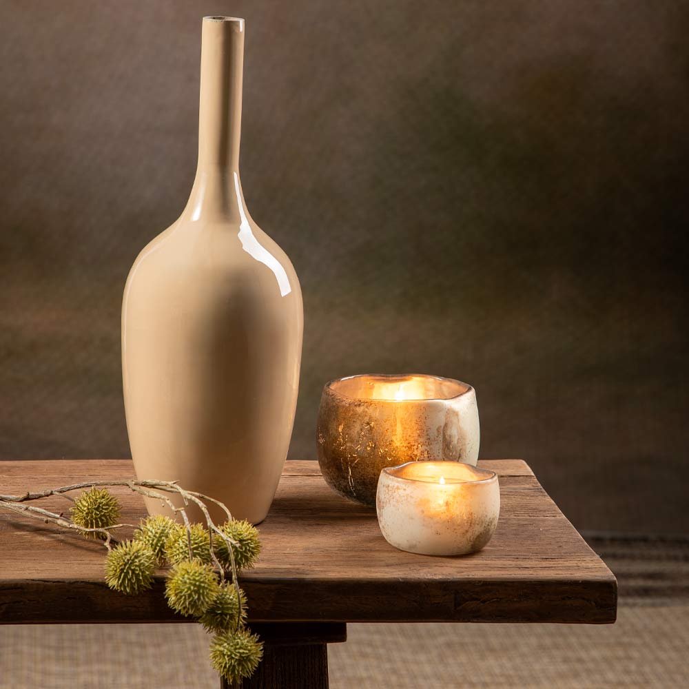 A tall, gloss beige metal vase stands on a rustic wooden table beside two lit candles in textured holders, with a sprig of faux green echinops in the foreground against a dark brown background wall.
