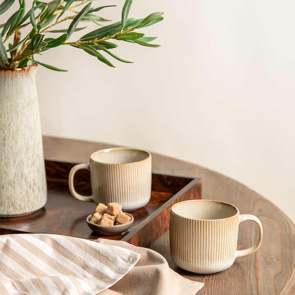 Two striped beige ceramic mugs with vertical ridges, one on a dark wooden tray and one on a wooden table, styled with a striped napkin, a small dish of sugar cubes, and a vase with green leaves against a warm off-white background.