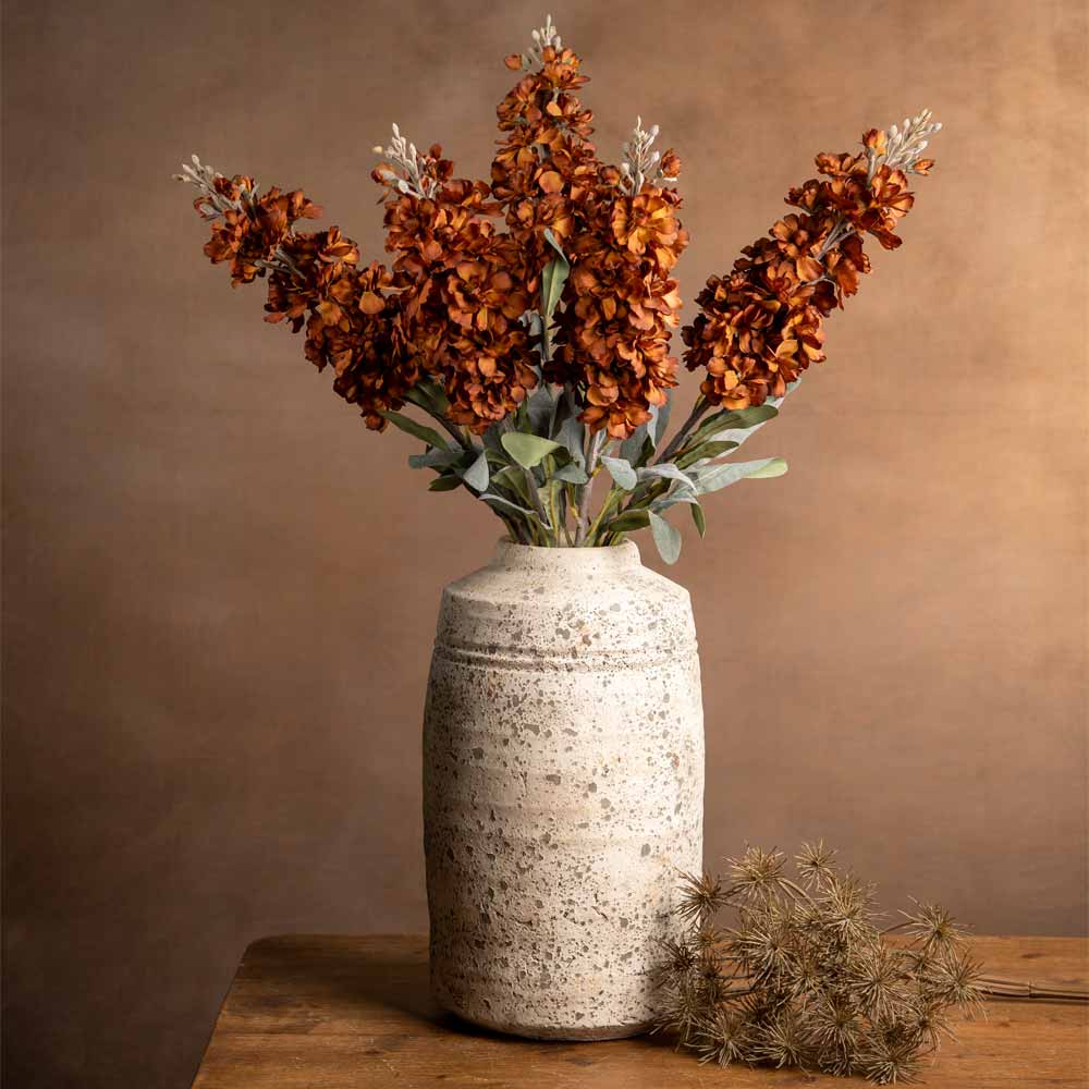 A rustic ceramic vase filled with Faux autumn Delphinium stems and green leaves, displayed on a wooden surface against a warm brown background.